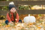 Little girl in beanie and vest sitting in leaves with white pumpkin