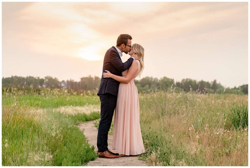 Regina Engagement Photography - Engagement Session - Couple kiss in a field of tall grass at sunset inside the Wascana Habitat Conservation Area - Blush chiffon dress - Black suit