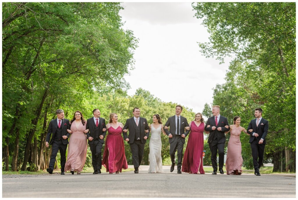 Taylor & Jolene - Emerald Park Wedding - 22 - Bridal party walks arm in arm down the road