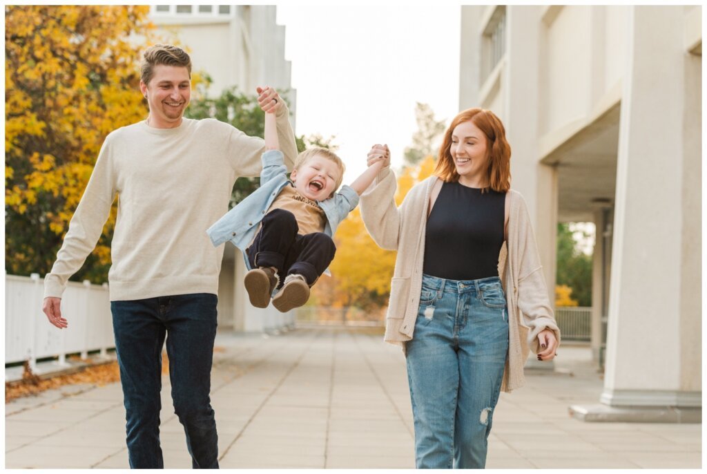 McFie Family - 05 - Regina Family Session Mom and dad swing their toddler by his hands