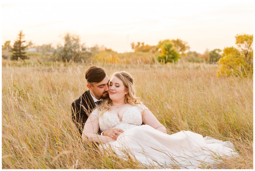 Orrin & Jade - 38 - Weyburn Wedding-Groom-holds-bride-as-they-sit-in-tall-grass