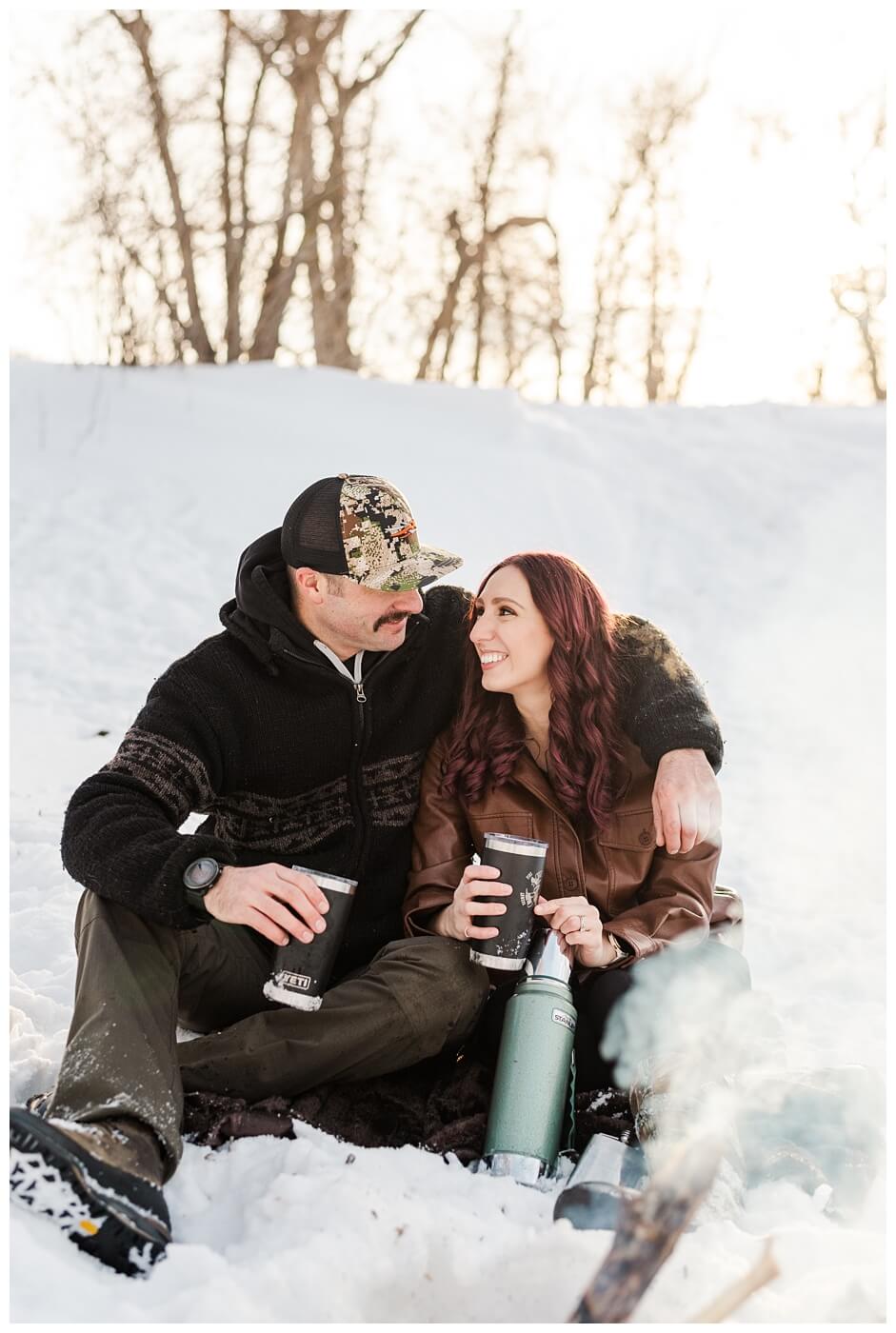 Brett & Brittany - Winter 2023 - Lumsden Valley - 01 - Couple sits drinking hot chocolate by the fire