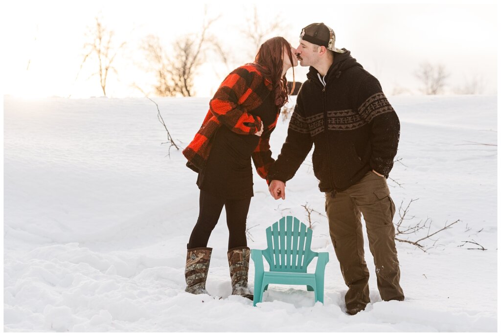 Brett & Brittany - Winter 2023 - Lumsden Valley - 11 - Husband & wife share a kiss overtop baby Adirondack chair