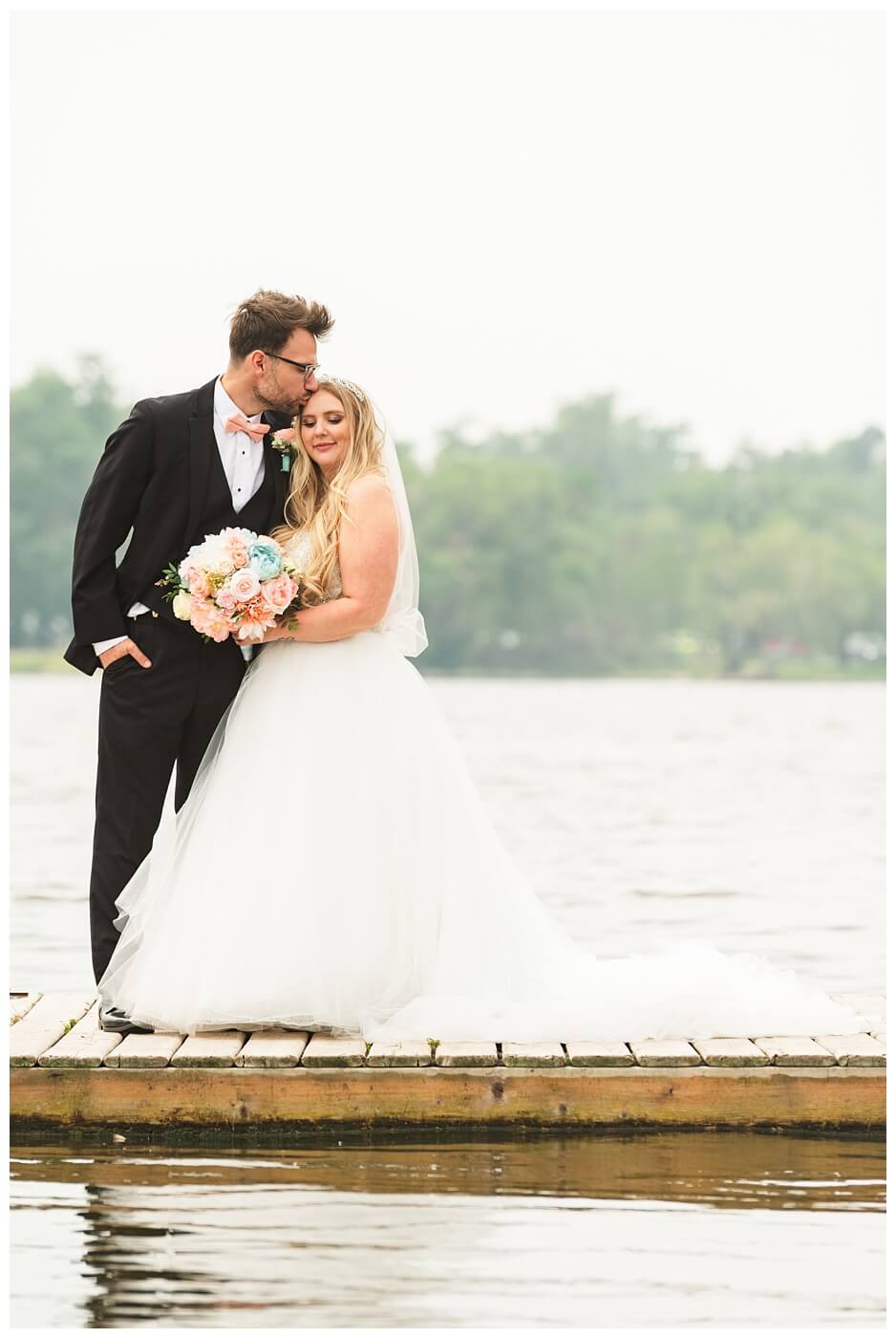 Brett & Rachelle - Delta Regina Wedding - 12 - Husband kissing wife's forehead on dock by Wascana