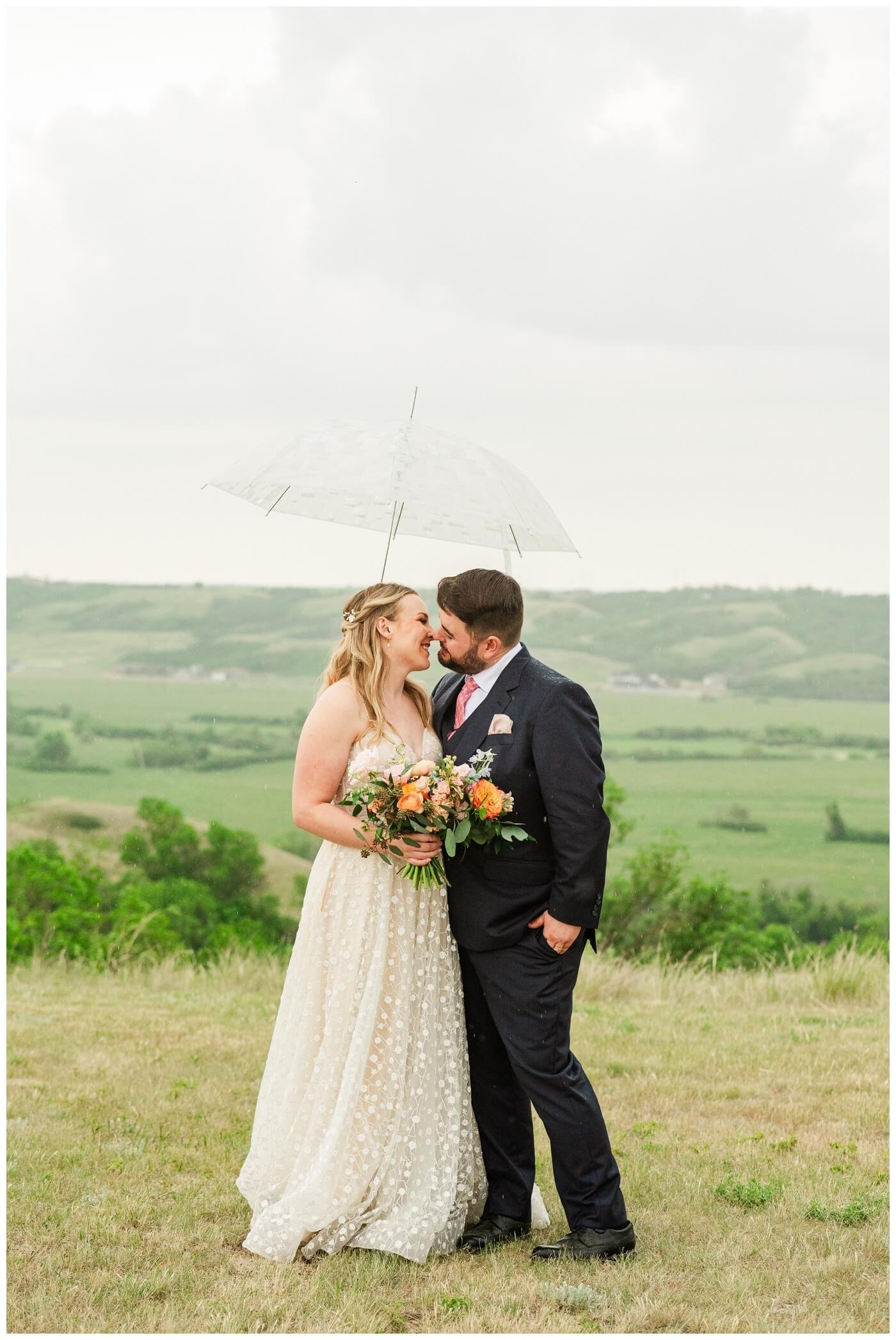 Darcy & Maddie - Regina Summer Wedding - 025 - Goodsman Farm - Bride and Groom under clear umbrella