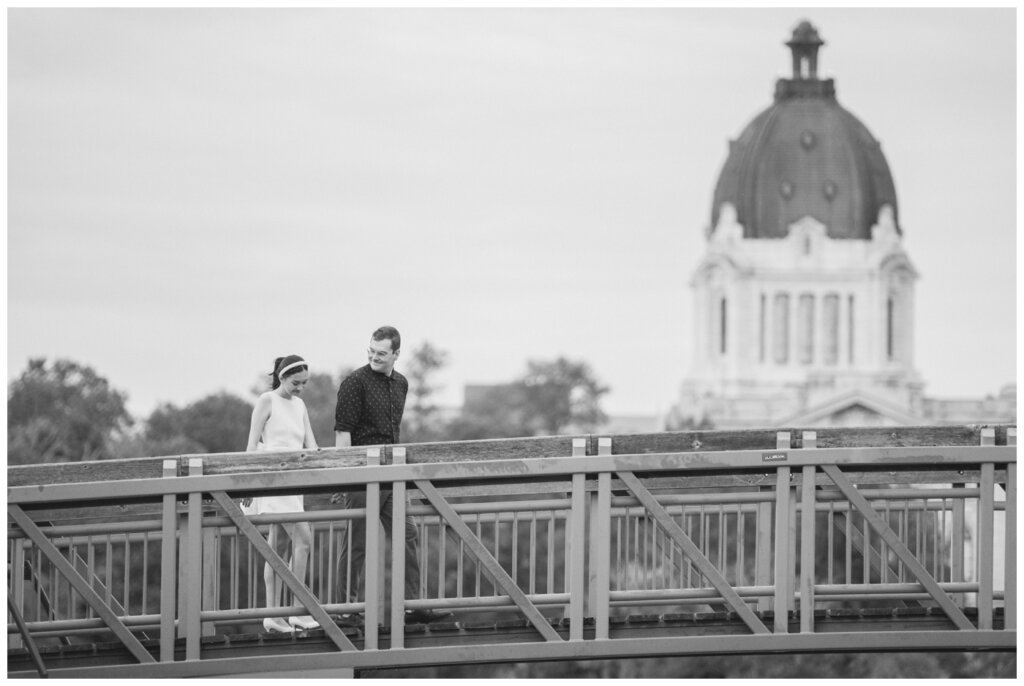 Skylar & Michaela- Regina Engagement - Wascana Park - 007 - Couple walk across bridge in front of the Legislative Building